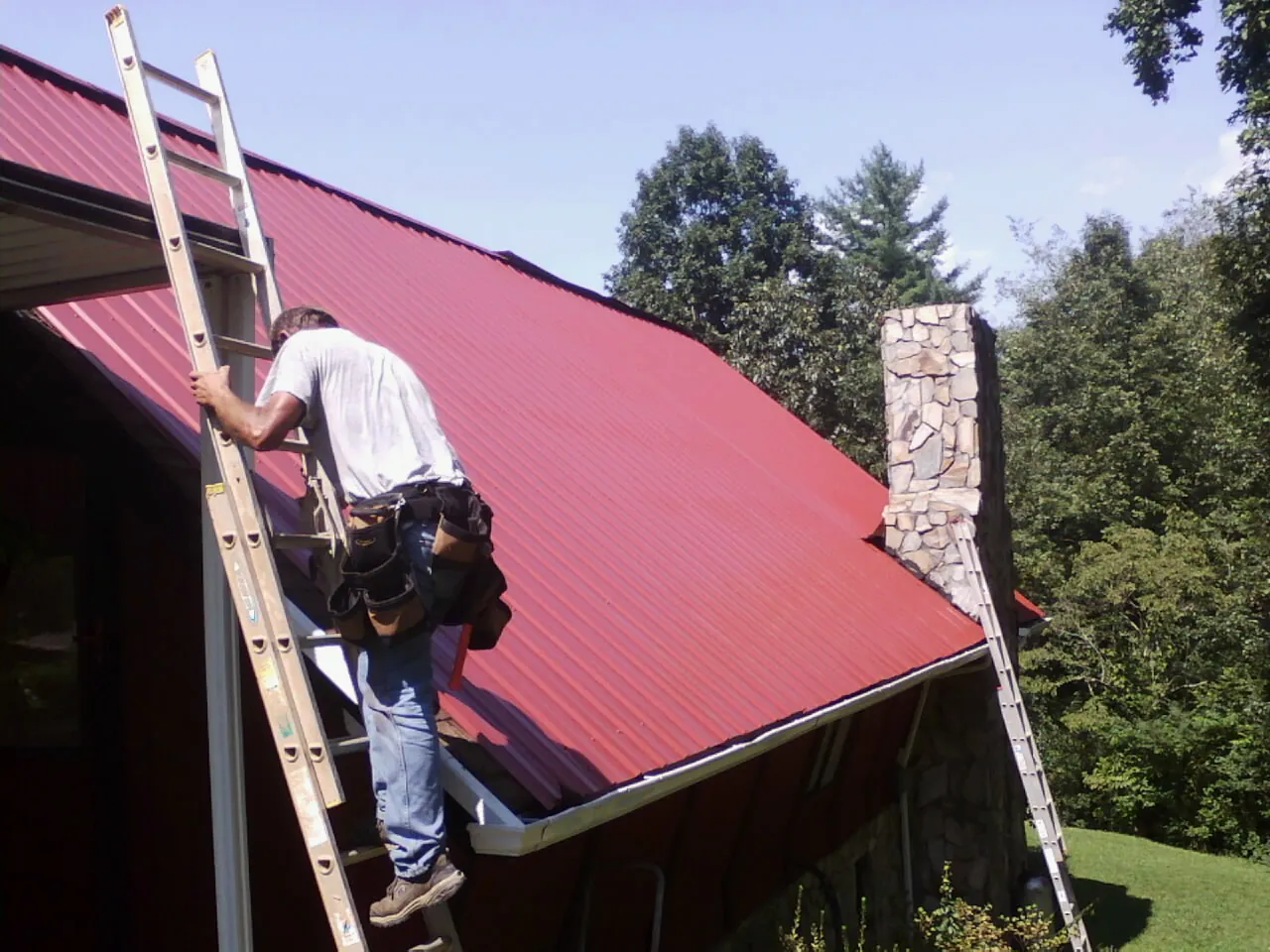Roofer on a ladder beside a red metal roof and stone chimney — Soffit & Fascia Repair by Roofing Brothers in Sussex